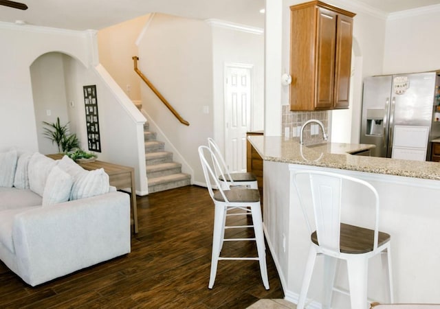 kitchen featuring light stone counters, dark wood-style flooring, crown molding, stainless steel refrigerator with ice dispenser, and brown cabinetry