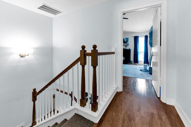 corridor featuring visible vents, a textured ceiling, an upstairs landing, wood finished floors, and baseboards