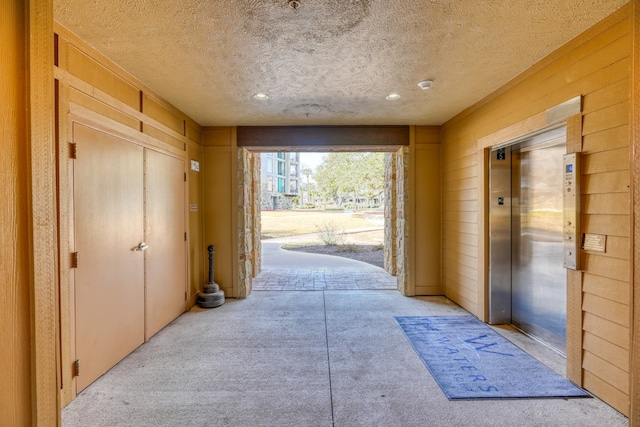 corridor featuring a textured ceiling, wooden walls, light colored carpet, and elevator