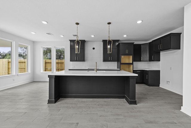 kitchen with light countertops, backsplash, a sink, an island with sink, and dark cabinets