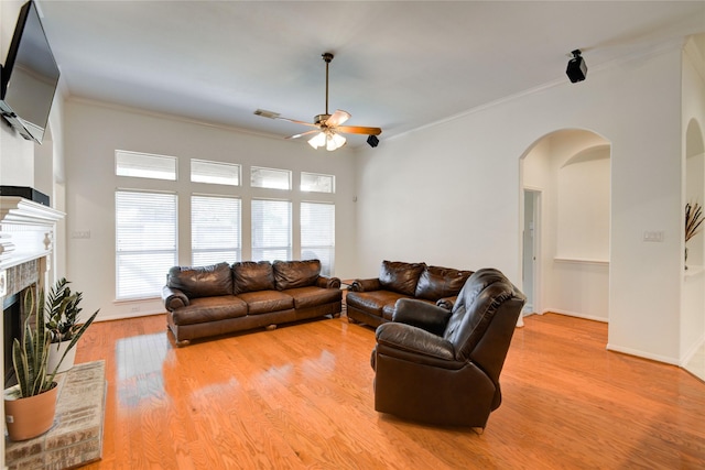 living room featuring arched walkways, a ceiling fan, light wood-style floors, crown molding, and a brick fireplace