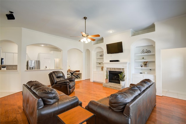 living room featuring built in shelves, wood finished floors, a ceiling fan, a brick fireplace, and crown molding