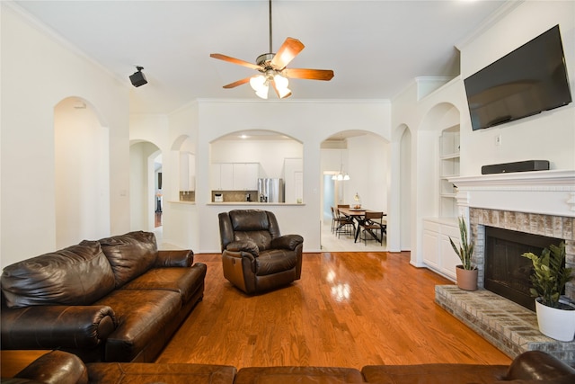 living room with built in shelves, a fireplace, crown molding, ceiling fan, and wood finished floors