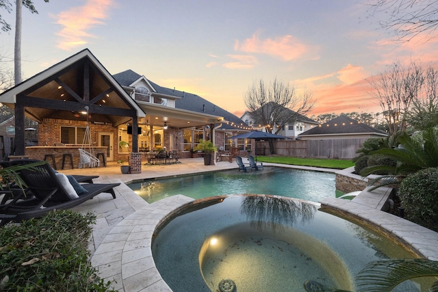 pool at dusk featuring a patio, ceiling fan, outdoor dry bar, fence, and a pool with connected hot tub