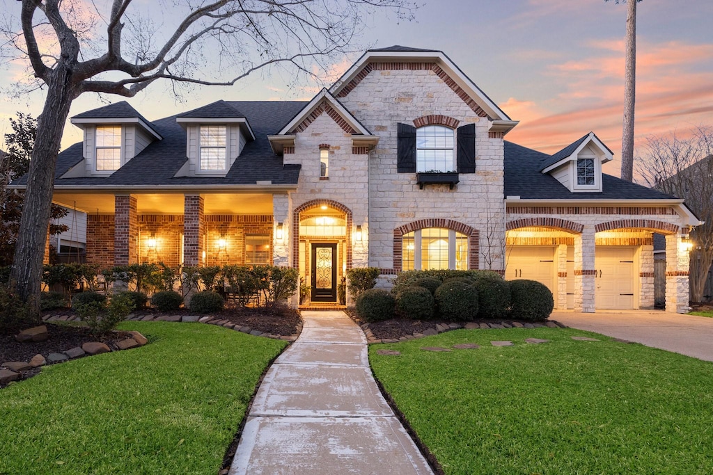 french country inspired facade featuring brick siding, a shingled roof, a front yard, stone siding, and driveway