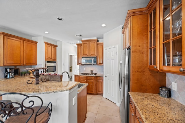 kitchen with appliances with stainless steel finishes, brown cabinetry, light tile patterned flooring, a sink, and light stone countertops