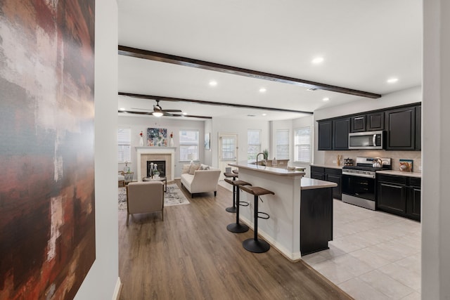 kitchen featuring a tiled fireplace, a kitchen bar, beam ceiling, stainless steel appliances, and dark cabinets