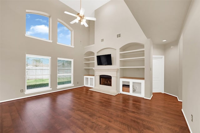 unfurnished living room featuring a fireplace with raised hearth, built in shelves, dark wood-type flooring, and plenty of natural light