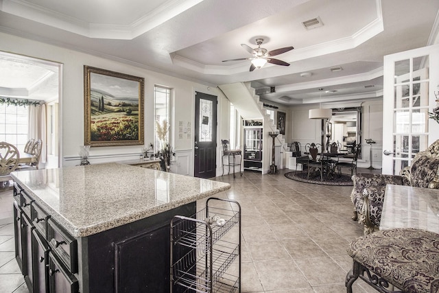 kitchen featuring wainscoting, light stone countertops, a tray ceiling, crown molding, and dark cabinetry