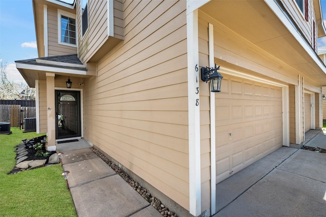 entrance to property featuring driveway, a garage, roof with shingles, fence, and central air condition unit