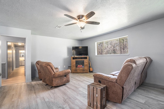 living area with a glass covered fireplace, light wood-style flooring, a ceiling fan, and visible vents