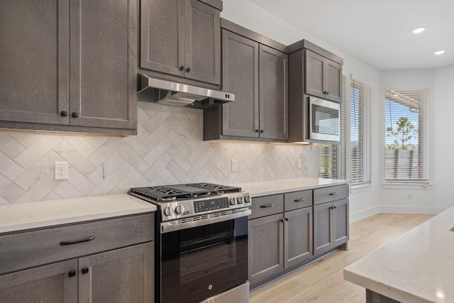 kitchen with baseboards, light wood-style floors, appliances with stainless steel finishes, under cabinet range hood, and backsplash