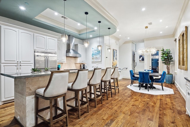 kitchen featuring built in fridge, wood finished floors, white cabinets, a kitchen breakfast bar, and wall chimney exhaust hood