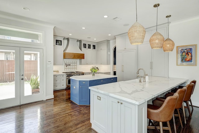 kitchen with custom range hood, high end stainless steel range oven, a kitchen island with sink, french doors, and blue cabinetry