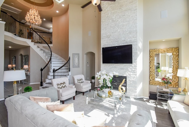 living area with arched walkways, a high ceiling, stairs, a stone fireplace, and ceiling fan with notable chandelier