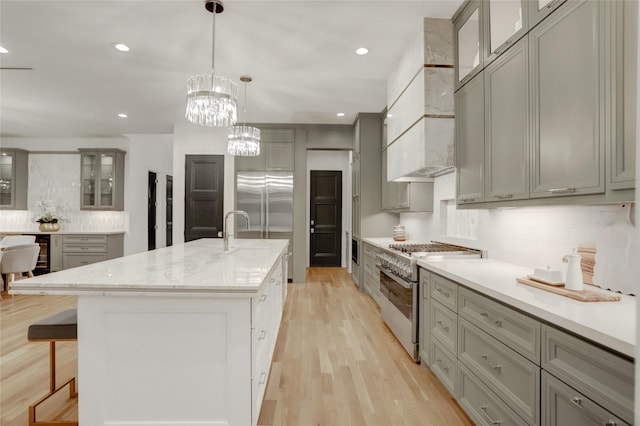 kitchen with an island with sink, high quality appliances, glass insert cabinets, light wood-type flooring, and premium range hood