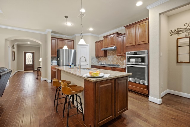 kitchen with arched walkways, stainless steel appliances, a sink, under cabinet range hood, and a kitchen bar