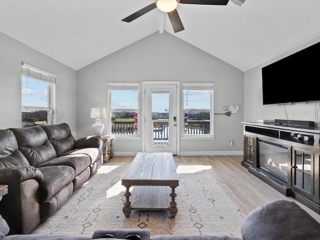 living area featuring baseboards, a ceiling fan, a glass covered fireplace, vaulted ceiling, and light wood-type flooring