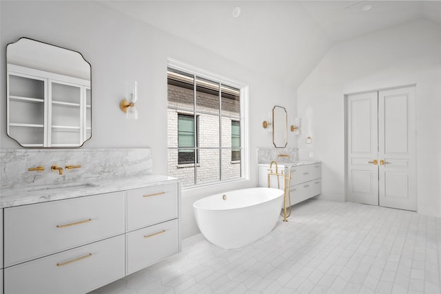 full bathroom with a soaking tub, two vanities, decorative backsplash, vaulted ceiling, and a sink