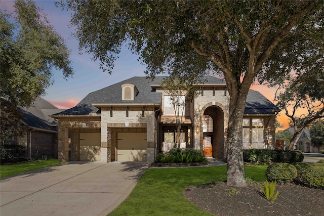 french country style house featuring an attached garage, a shingled roof, a lawn, stone siding, and driveway