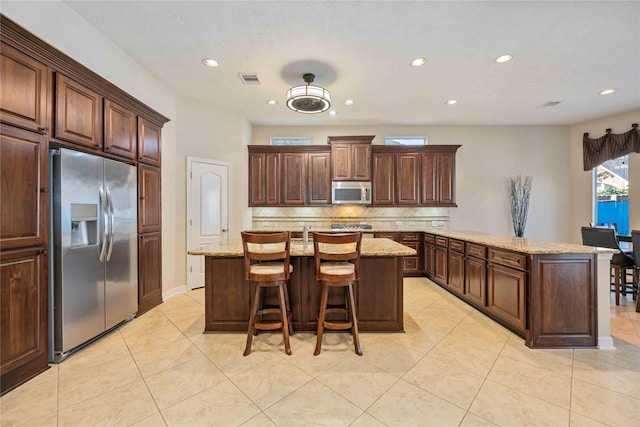 kitchen with an island with sink, light stone counters, a peninsula, appliances with stainless steel finishes, and decorative backsplash