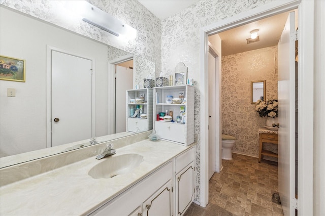 bathroom with stone finish floor, vanity, toilet, and wallpapered walls