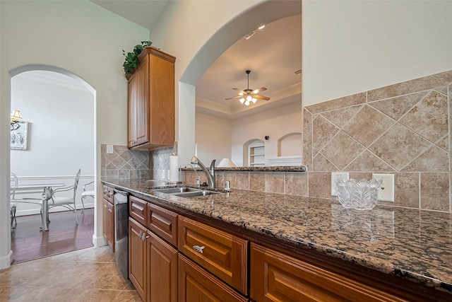 kitchen featuring tasteful backsplash, dark stone counters, a tray ceiling, a ceiling fan, and a sink