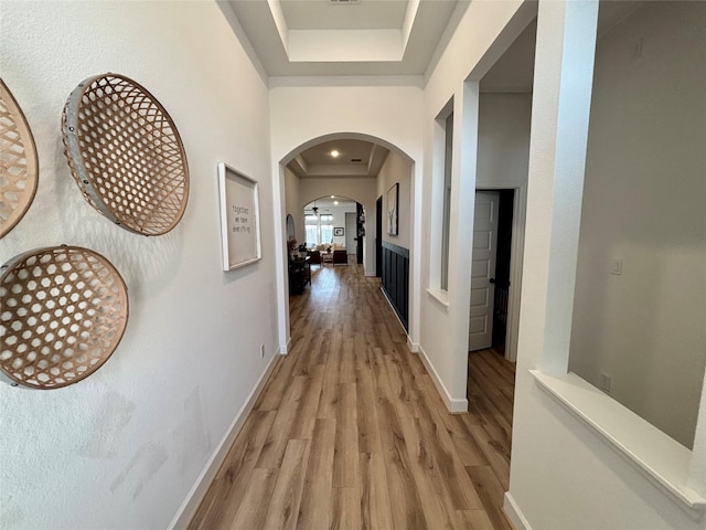 hallway featuring light wood-style floors, arched walkways, baseboards, and a tray ceiling