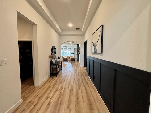 hall featuring light wood finished floors, visible vents, arched walkways, a raised ceiling, and baseboards