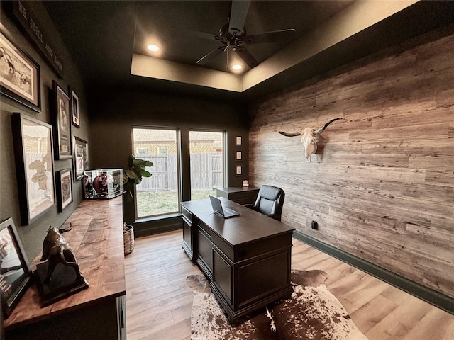 home office with ceiling fan, light wood-style flooring, wood walls, a fireplace, and a raised ceiling