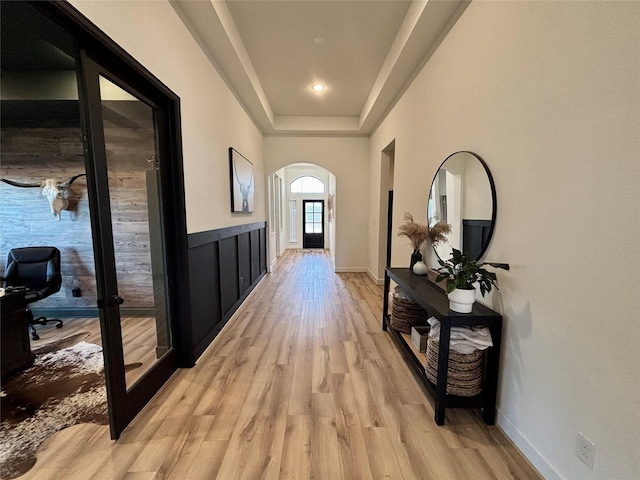 corridor with arched walkways, light wood finished floors, a raised ceiling, and baseboards