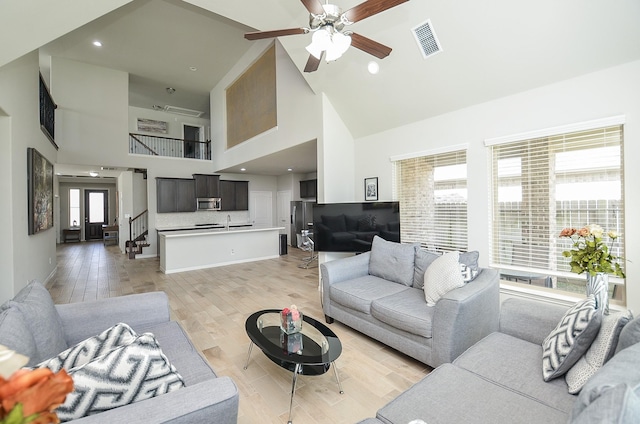 living area featuring ceiling fan, high vaulted ceiling, light wood-style flooring, visible vents, and stairway