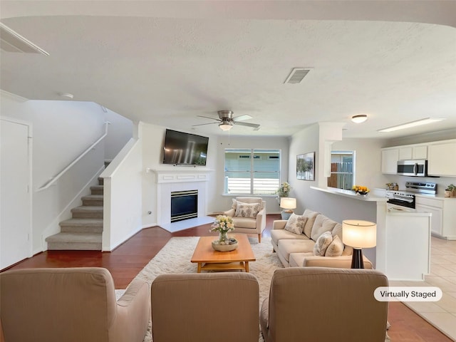 living room featuring a ceiling fan, visible vents, a fireplace with flush hearth, stairs, and a textured ceiling