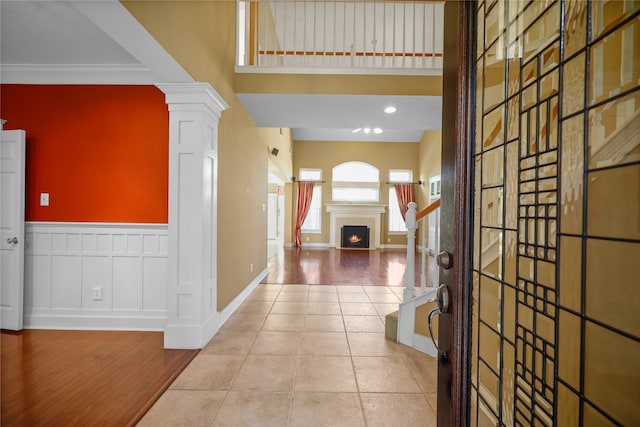 tiled entrance foyer featuring decorative columns, a warm lit fireplace, ornamental molding, a towering ceiling, and wainscoting