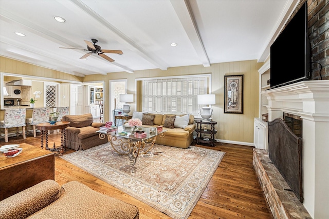 living room featuring beam ceiling, a fireplace with raised hearth, a ceiling fan, wood finished floors, and baseboards