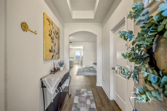 corridor featuring a tray ceiling, arched walkways, dark wood-type flooring, and baseboards