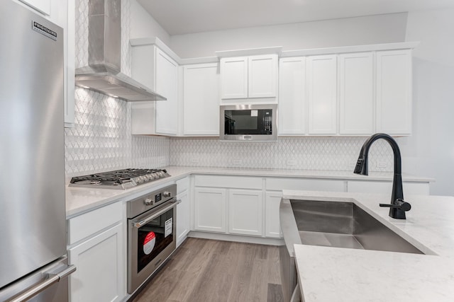 kitchen featuring stainless steel appliances, a sink, light wood-style floors, wall chimney range hood, and decorative backsplash