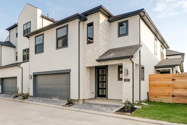 multi unit property featuring a shingled roof, stone siding, an attached garage, fence, and stucco siding