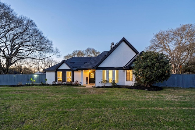 back of house at dusk featuring fence, a yard, a chimney, brick siding, and roof mounted solar panels