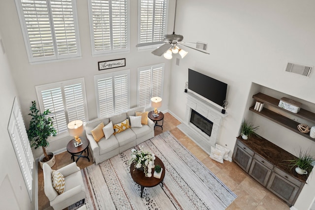 living room featuring baseboards, visible vents, a ceiling fan, a glass covered fireplace, and a high ceiling