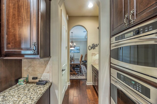 kitchen featuring backsplash, dark brown cabinets, dark wood-type flooring, light stone countertops, and double oven