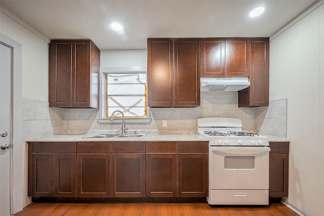 kitchen with white gas range oven, wood finished floors, a sink, under cabinet range hood, and backsplash