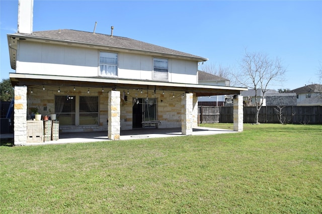 back of property featuring fence, stone siding, a yard, roof with shingles, and a patio area