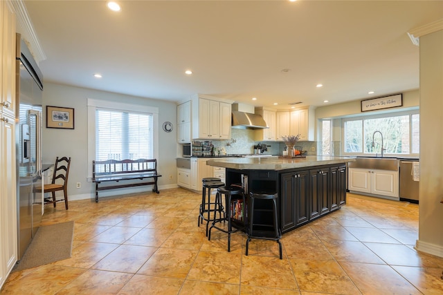 kitchen with backsplash, appliances with stainless steel finishes, a sink, a kitchen island, and wall chimney exhaust hood