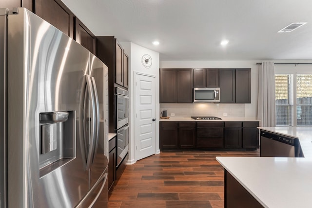 kitchen with dark brown cabinetry, stainless steel appliances, dark wood-type flooring, light countertops, and tasteful backsplash