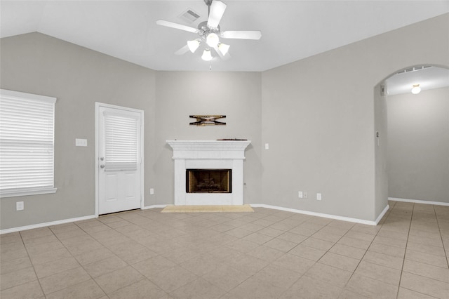 unfurnished living room featuring a fireplace with raised hearth, baseboards, ceiling fan, light tile patterned floors, and arched walkways