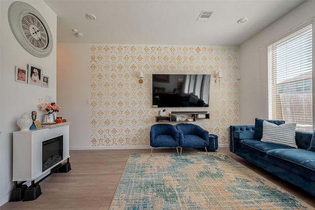 living area featuring visible vents, wood finished floors, a glass covered fireplace, wallpapered walls, and baseboards