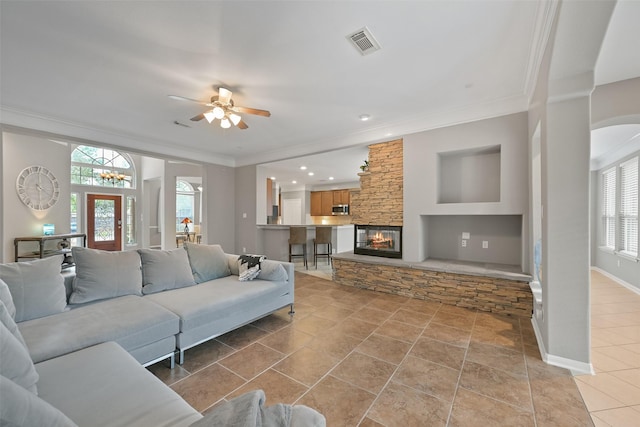 living area featuring baseboards, visible vents, a ceiling fan, ornamental molding, and a stone fireplace