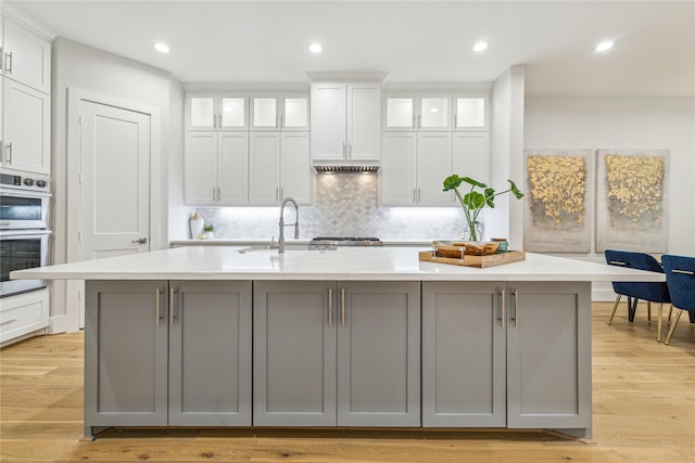 kitchen with stainless steel double oven, gray cabinetry, white cabinetry, light countertops, and an island with sink