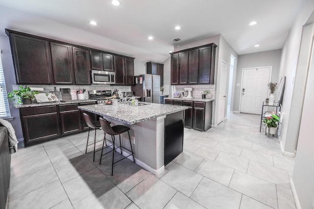 kitchen with a center island with sink, visible vents, appliances with stainless steel finishes, dark brown cabinetry, and light stone countertops